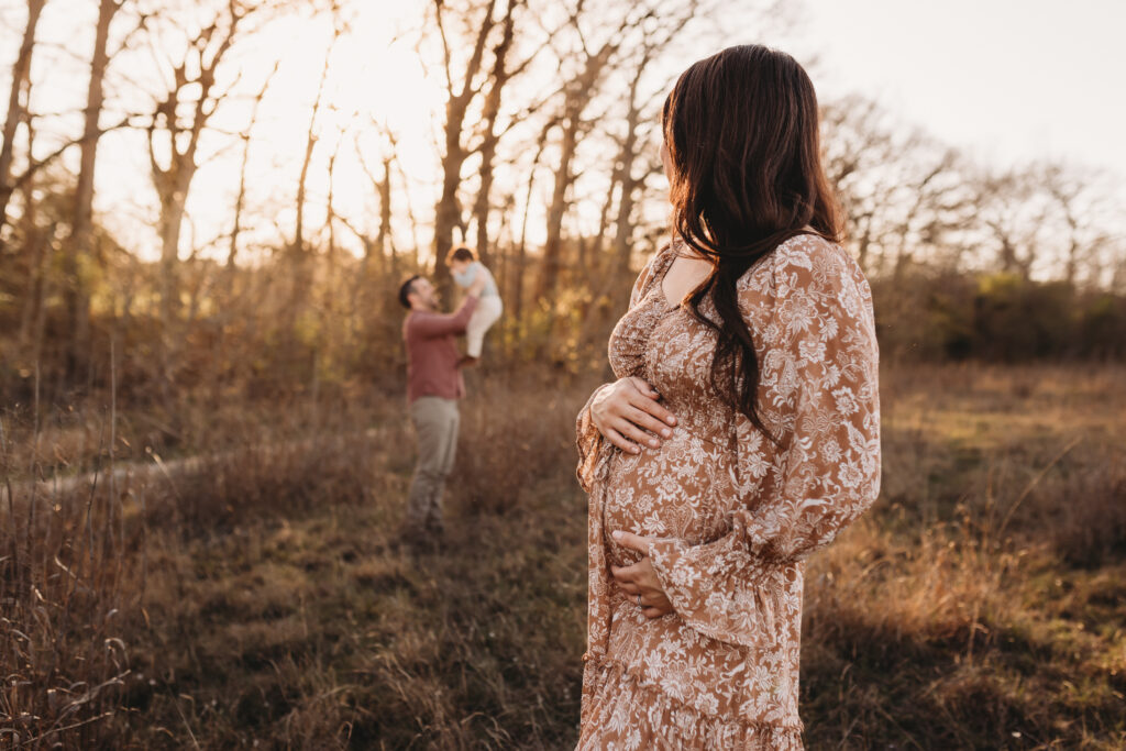 pregnant woman holding her belly while looking at husband and son playing in the background at Cibolo nature Center